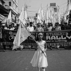 A young girl in India at the front of a student rally for economic and social justice, holding a flag with a banner behind her.