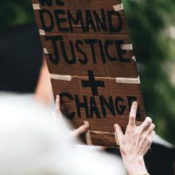 A protest sign advocating for social transformation. The sign reads "We Demand Justice + Change".