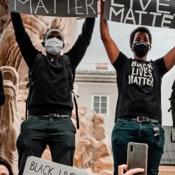 An image of a Black Lives Matter protest taking place by a fountain. People are holding signs reading 'Black Lives Matter!', 'Time to Change the World' and 'Wir stehen auf geggen rassismus'.