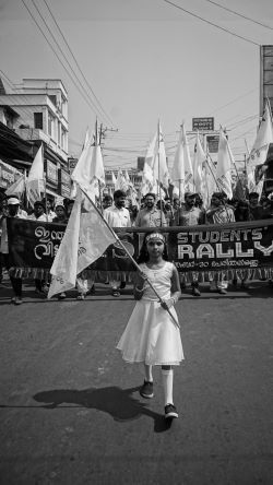 A young girl in India at the front of a student rally for economic and social justice, holding a flag with a banner behind her.