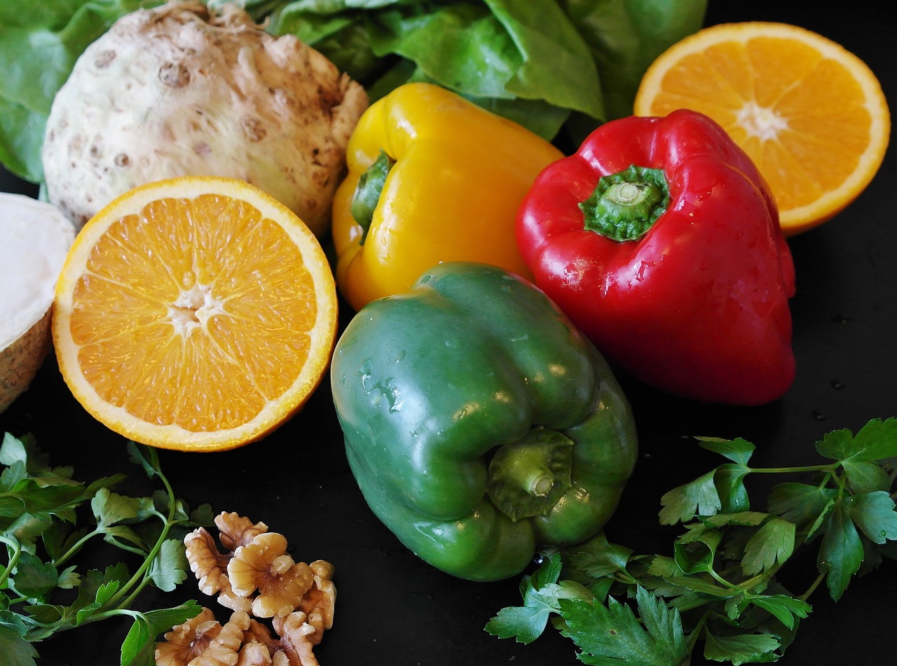 Fruits and vegetables- key parts of a healthier lifestyle. A selection of fruit and vegetables arranged on a table, including a cut-open orange, red, yellow, and green peppers, a fennel bulb and walnuts. All examples of what we can consume to achieve healthier diets.