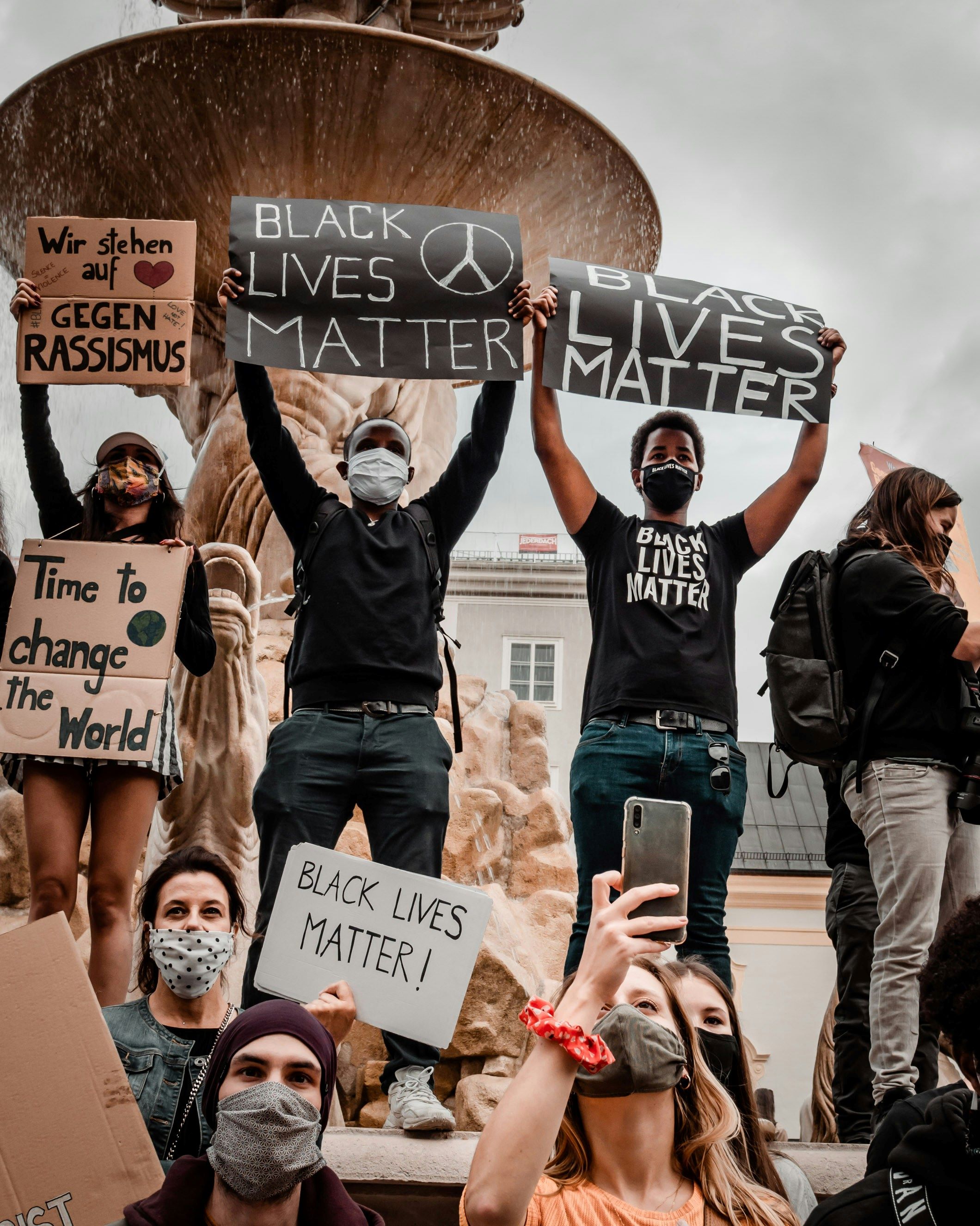 An image of a Black Lives Matter protest taking place by a fountain. People are holding signs reading 'Black Lives Matter!', 'Time to Change the World' and 'Wir stehen auf geggen rassismus'.